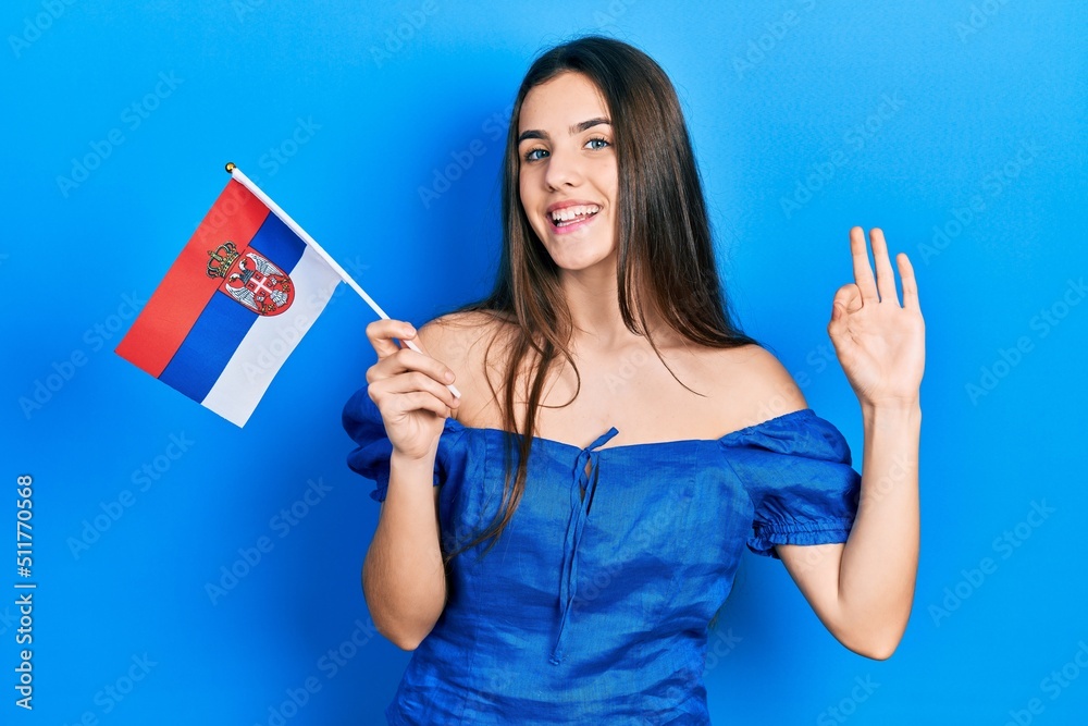 Young brunette teenager holding serbia flag doing ok sign with fingers, smiling friendly gesturing excellent symbol Young brunette teenager holding serbia flag doing ok sign with fingers, smiling friendly gesturing excellent symbol