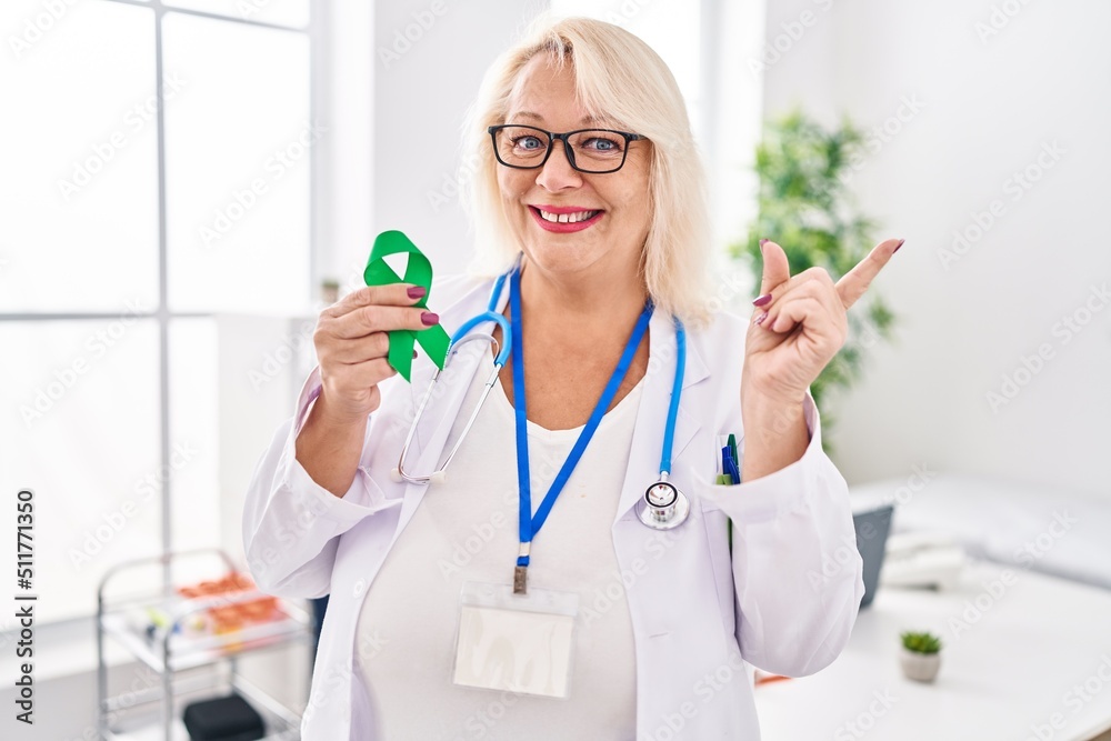 Middle age caucasian woman holding support green ribbon smiling happy pointing with hand and finger to the side Middle age caucasian woman holding support green ribbon smiling happy pointing with hand and finger to the side