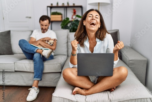 Hispanic middle age couple at home, woman using laptop very happy and excited doing winner gesture with arms raised, smiling and screaming for success. celebration concept. © Krakenimages.com