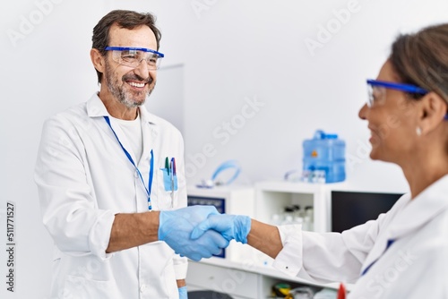 Middle age man and woman partners wearing scientist uniform shake hands at laboratory