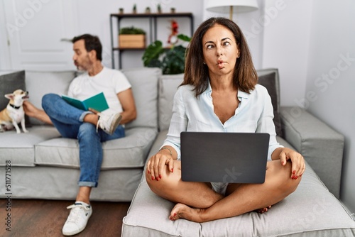 Hispanic middle age couple at home, woman using laptop making fish face with lips, crazy and comical gesture. funny expression. © Krakenimages.com