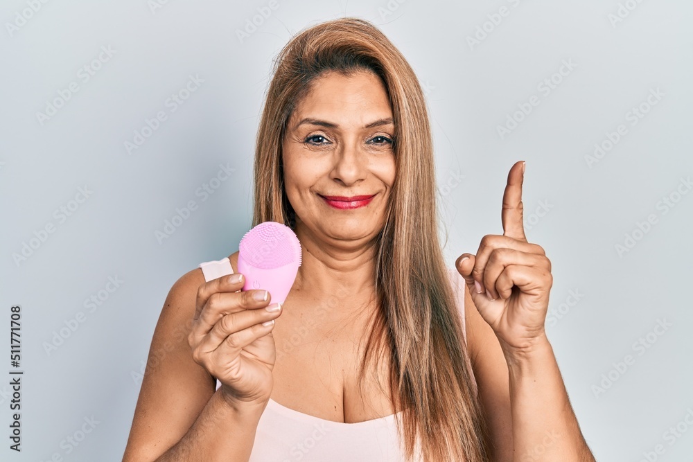 Middle age hispanic woman using facial exfoliating cleaner smiling with an idea or question pointing finger with happy face, number one
