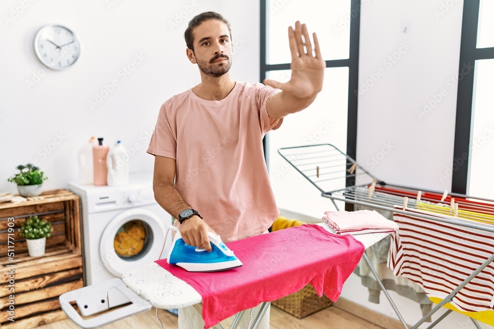 Obraz premium Young hispanic man ironing clothes at home doing stop sing with palm of the hand. warning expression with negative and serious gesture on the face.