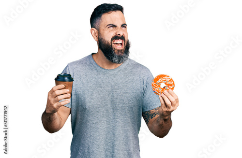 Hispanic man with beard eating doughnut and drinking coffee angry and mad screaming frustrated and furious, shouting with anger looking up.
