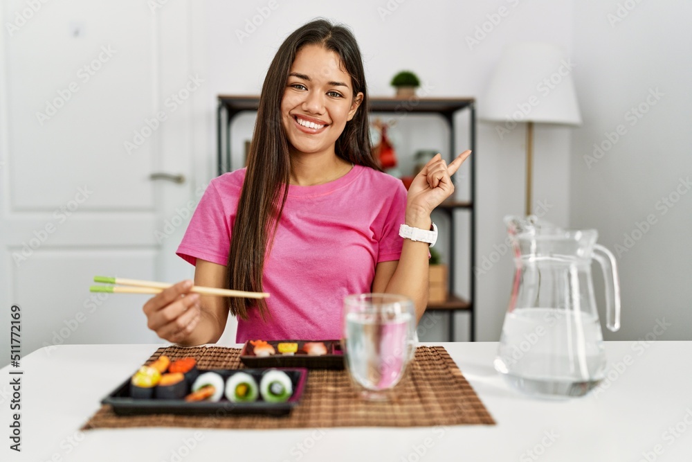 Young brunette woman eating sushi using chopsticks with a big smile on face, pointing with hand finger to the side looking at the camera.
