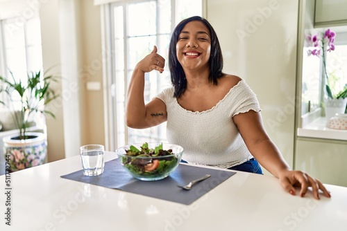 Young hispanic woman eating healthy salad at home smiling doing phone gesture with hand and fingers like talking on the telephone. communicating concepts.