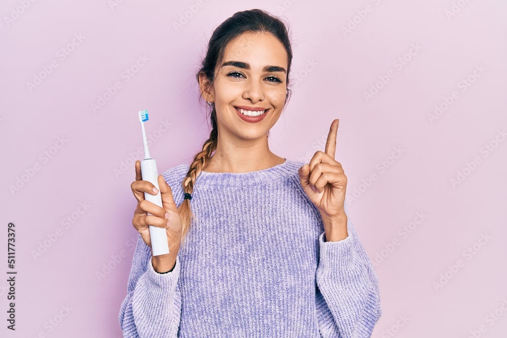 Young hispanic girl holding electric toothbrush smiling with an idea or question pointing finger with happy face, number one Young hispanic girl holding electric toothbrush smiling with an idea or question pointing finger with happy face, number one