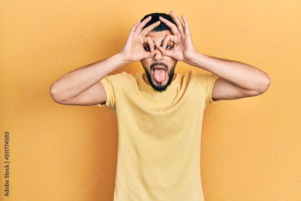 Handsome man with beard wearing casual yellow t shirt doing ok gesture like binoculars sticking tongue out, eyes looking through fingers. crazy expression. Handsome man with beard wearing casual yellow t shirt doing ok gesture like binoculars sticking tongue out, eyes looking through fingers. crazy expression.