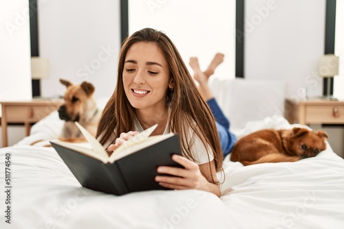 Young hispanic woman reading book lying on bed with dogs at bedroom