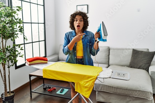 Young hispanic woman ironing clothes at home surprised pointing with finger to the side, open mouth amazed expression.