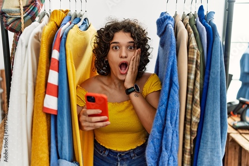 Young hispanic woman searching clothes on clothing rack using smartphone afraid and shocked, surprise and amazed expression with hands on face