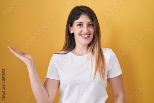 Young brunette woman standing over yellow background smiling cheerful presenting and pointing with palm of hand looking at the camera.