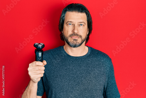 Middle age caucasian man holding electric razor machine thinking attitude and sober expression looking self confident