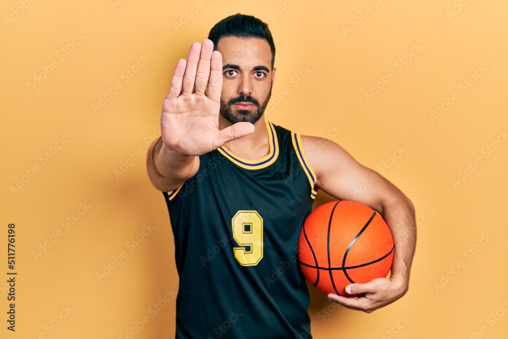 Handsome hispanic man with beard holding basketball ball with open hand doing stop sign with serious and confident expression, defense gesture