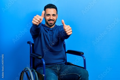 Handsome hispanic man with beard sitting on wheelchair approving doing positive gesture with hand, thumbs up smiling and happy for success. winner gesture.