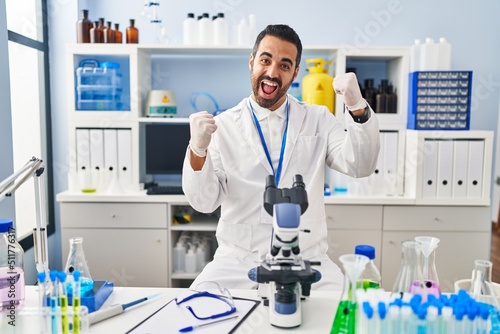 Young hispanic man with beard working at scientist laboratory very happy and excited doing winner gesture with arms raised, smiling and screaming for success. celebration concept.
