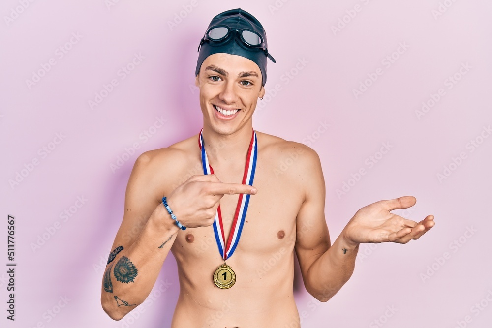Young hispanic man wearing swimmer glasses and gold medal amazed and smiling to the camera while presenting with hand and pointing with finger. Young hispanic man wearing swimmer glasses and gold medal amazed and smiling to the camera while presenting with hand and pointing with finger.