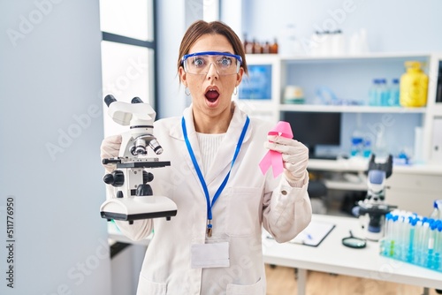 Young brunette woman working at scientist laboratory holding pink ribbon in shock face, looking skeptical and sarcastic, surprised with open mouth