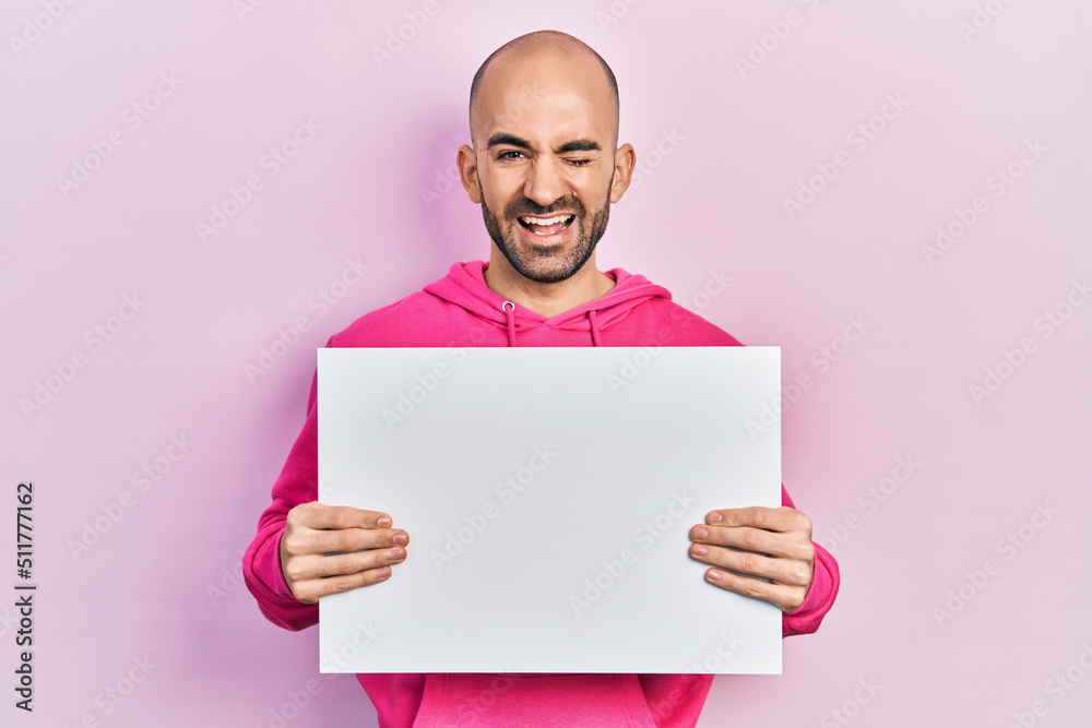 Young bald man holding blank empty banner winking looking at the camera with sexy expression, cheerful and happy face. Young bald man holding blank empty banner winking looking at the camera with sexy expression, cheerful and happy face.