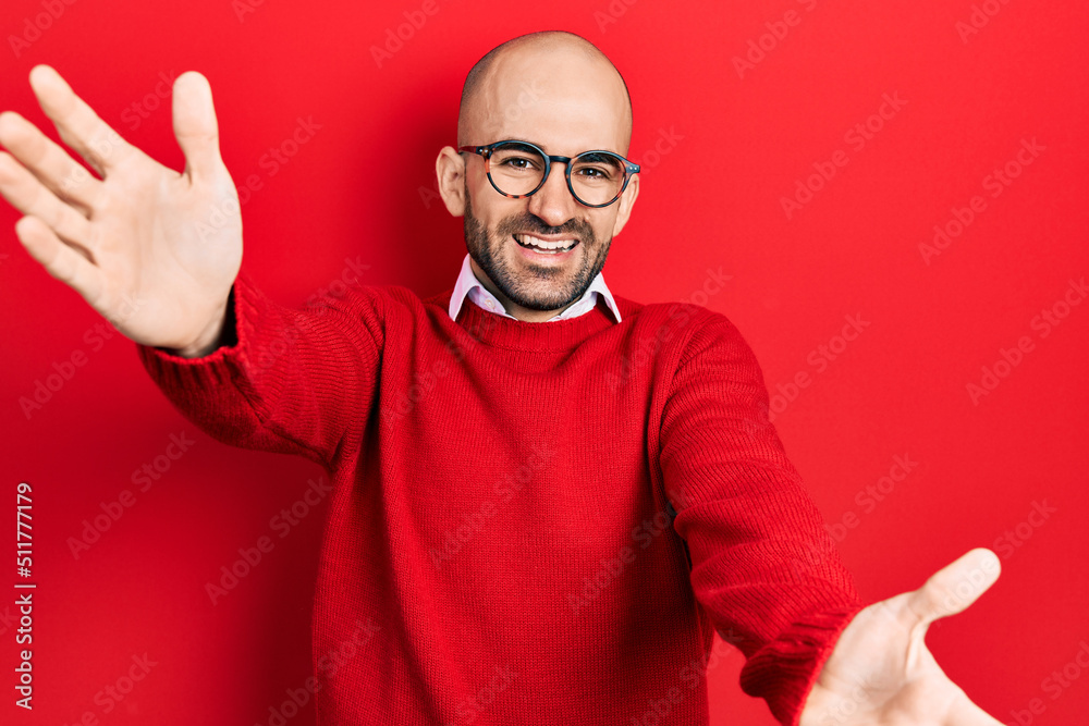 Naklejka premium Young bald man wearing casual clothes and glasses looking at the camera smiling with open arms for hug. cheerful expression embracing happiness.