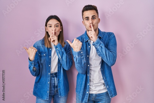 Young hispanic couple standing over pink background asking to be quiet with finger on lips pointing with hand to the side. silence and secret concept.