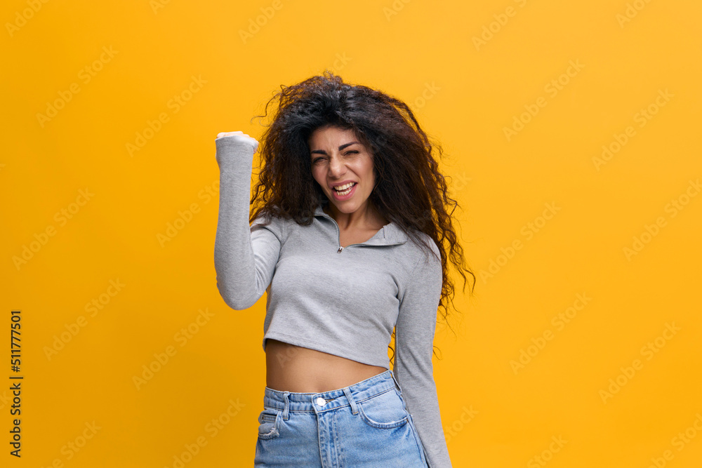 Excited amazed young curly african american woman 20s wearing casual gray sweatshirt standing doing winner gesture clenching fist isolated on pastel yellow color background studio portrait Excited amazed young curly african american woman 20s wearing casual gray sweatshirt standing doing winner gesture clenching fist isolated on pastel yellow color background studio portrait