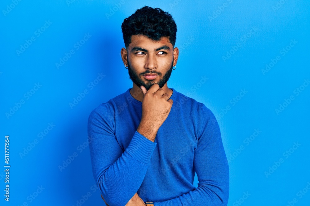 Arab man with beard wearing casual blue sweater with hand on chin thinking about question, pensive expression. smiling with thoughtful face. doubt concept. Arab man with beard wearing casual blue sweater with hand on chin thinking about question, pensive expression. smiling with thoughtful face. doubt concept.