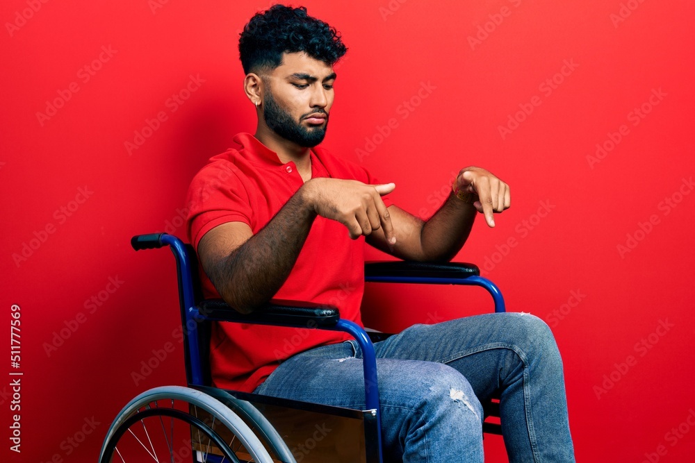 Arab man with beard sitting on wheelchair pointing down looking sad and upset, indicating direction with fingers, unhappy and depressed. Arab man with beard sitting on wheelchair pointing down looking sad and upset, indicating direction with fingers, unhappy and depressed.