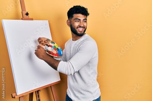 Fotografiet Arab man with beard standing drawing with palette by painter easel stand smiling looking to the side and staring away thinking