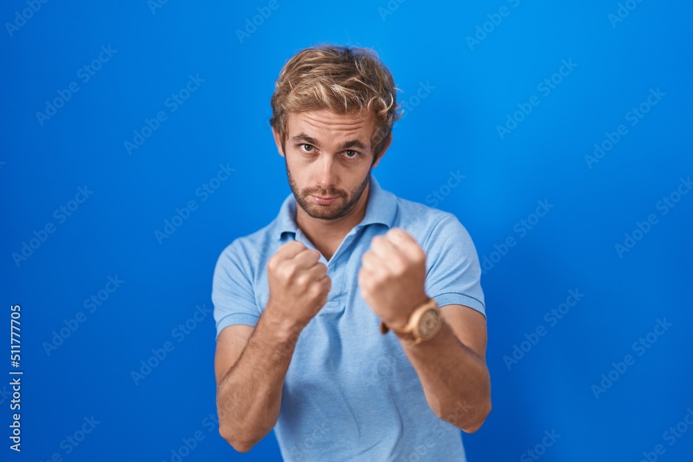 Caucasian man standing over blue background ready to fight with fist defense gesture, angry and upset face, afraid of problem Caucasian man standing over blue background ready to fight with fist defense gesture, angry and upset face, afraid of problem