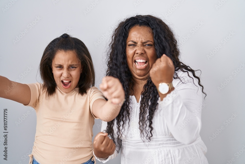 Mother and young daughter standing over white background angry and mad raising fists frustrated and furious while shouting with anger. rage and aggressive concept.