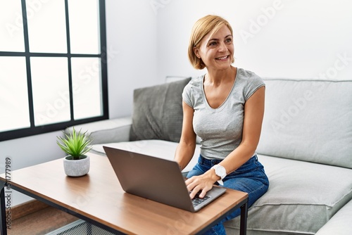 Middle age blonde woman using laptop at home looking away to side with smile on face  natural expression. laughing confident.