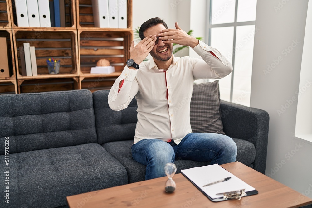 Young hispanic man with beard working at consultation office covering eyes with hands smiling cheerful and funny. blind concept.