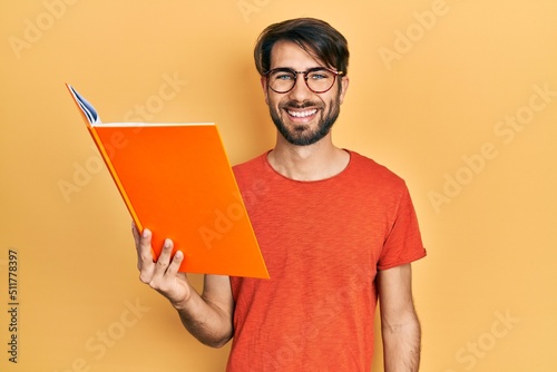 Young hispanic man reading book looking positive and happy standing and smiling with a confident smile showing teeth
