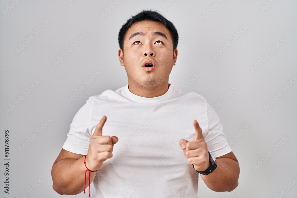 Young chinese man standing over white background amazed and surprised looking up and pointing with fingers and raised arms. Young chinese man standing over white background amazed and surprised looking up and pointing with fingers and raised arms.