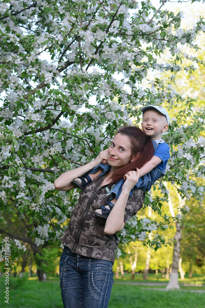 A young mother with her little son are walking in the spring garden. A little boy sits on his mother's neck. A young mother with her little son are walking in the spring garden. A little boy sits on his mother's neck.