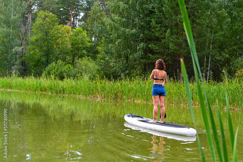 A woman drives on the Sup Board through a narrow canal surrounded by dense grass. Active weekend vacations wild nature outdoor. The woman is standing in a jeans.
