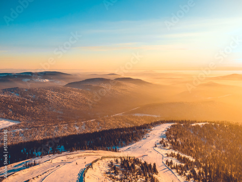 Ski slope in a mountainous area in the forest at sunset, aerial view. Skiers and snowboarders roll down the mountain with stunning views of nature.