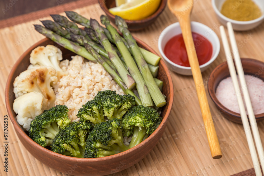 Baked broccoli, cauliflower and asparagus with brown rice, spices and lemon in clay bowl with chopsticks. Vegan vegetarian healthy asian food