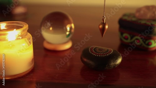 Golden color pendulum swinging on top of painted black stone with crystal bowl and blue box on wood table with orange light photo