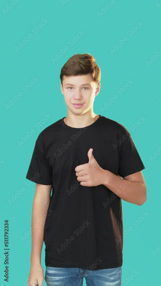 Like gesture. Good job. Approval sign. Support encourage. Smiling teenager boy showing thumbs up bowing head looking at camera isolated turquoise vertical. Like gesture. Good job. Approval sign. Support encourage. Smiling teenager boy showing thumbs up bowing head looking at camera isolated turquoise vertical.