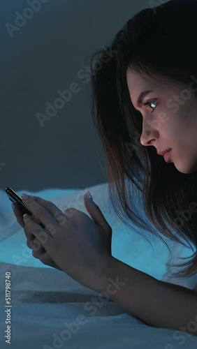 Online communication. Sleepless night. Virtual life. Pretty smiling woman laying at home bed scrolling smartphone in light shadow room interior. Vertical video.