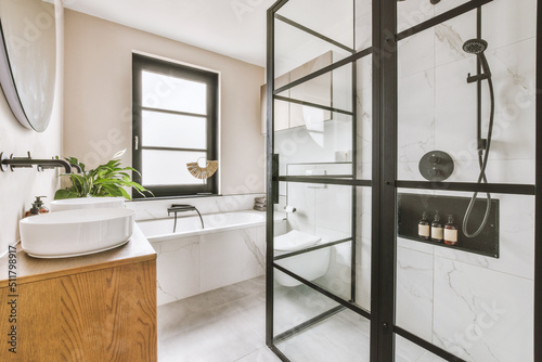 Shower faucets attached to tiled wall near glass partition, toilet, sink and ornamental curtail in washroom at home