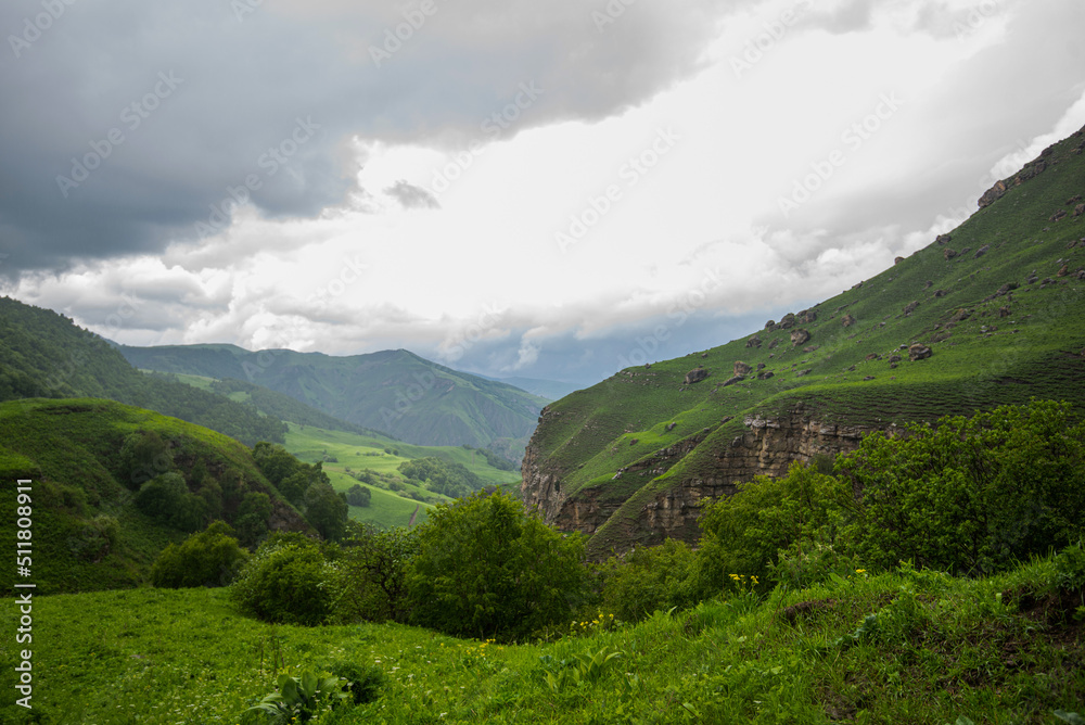 Beautiful spring landscape in the Caucasus Mountains with lush grassy hills and rocky slopes. Dramatic cloudy sky on a rainy day. Low clouds on the tops of the mountains.