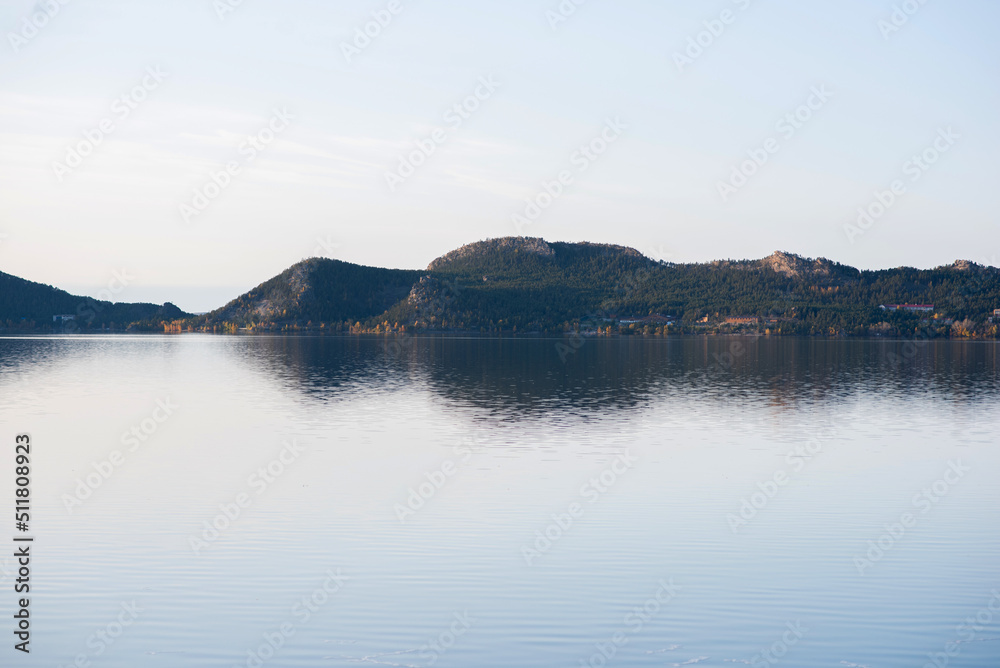 A lake with morning haze, calm water and reflection. Idyllic lake view. There are mountain ranges around the lake. The calm surface of the lake. There is a dense forest on the shore.