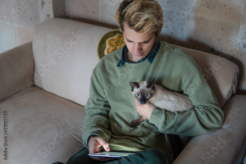 Man carrying his Siamese cat in the living room of his house while working on the sofa with his tablet