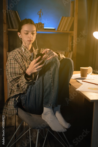 Relaxed asian woman student wearing headphones watching media on smart phone in the night at home, girl listens music with earphones on phone