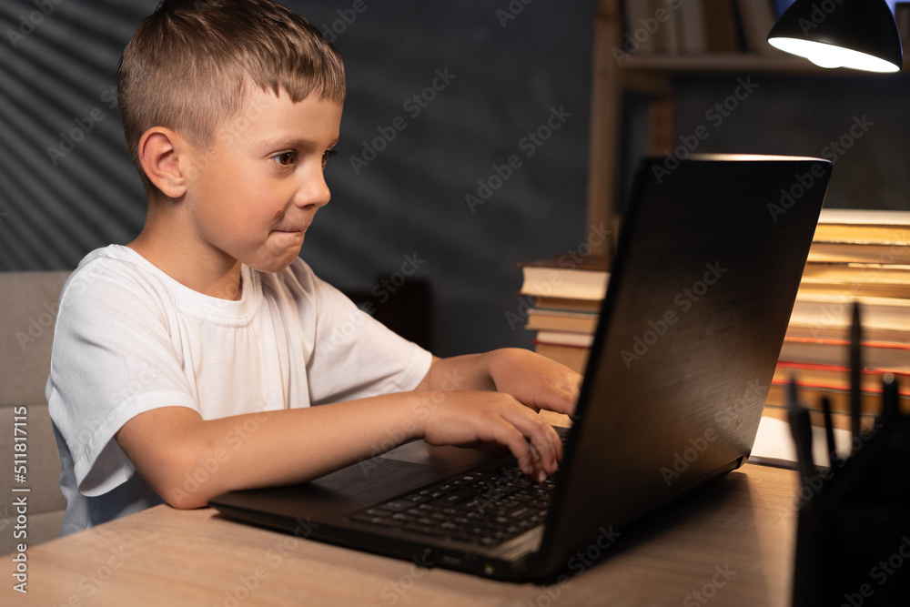 Portrait of child schoolboy studying at home using laptop computer at night, teenager with laptop doing homework at table in evening