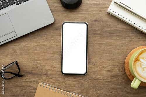 Smartphone with blank empty mockup screen is on top of wood office desk table with laptop computer and supplies. Top view with copy space, flat lay.