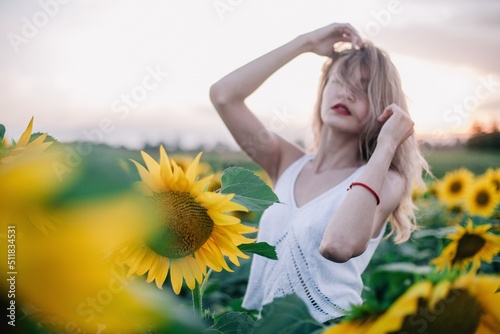 A young, slender girl with loose hair in a T-shirt stands in a field of sunflowers at sunset © Denis